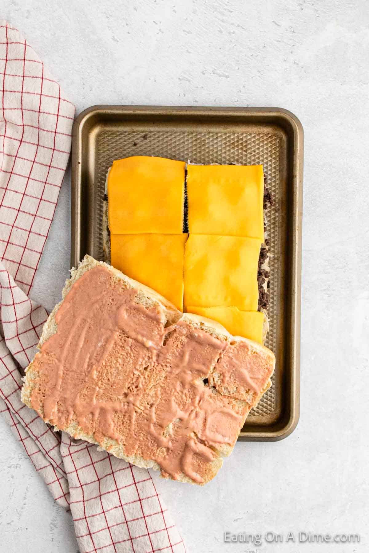 A baking tray with a rectangular sandwich. The bottom half has cooked ground beef topped with square slices of melted yellow cheese. The top half, harkening to a Hawaiian Roll Hamburger recipe, is spread with a pinkish sauce and placed upside down. A red and white checkered cloth is beside the tray.