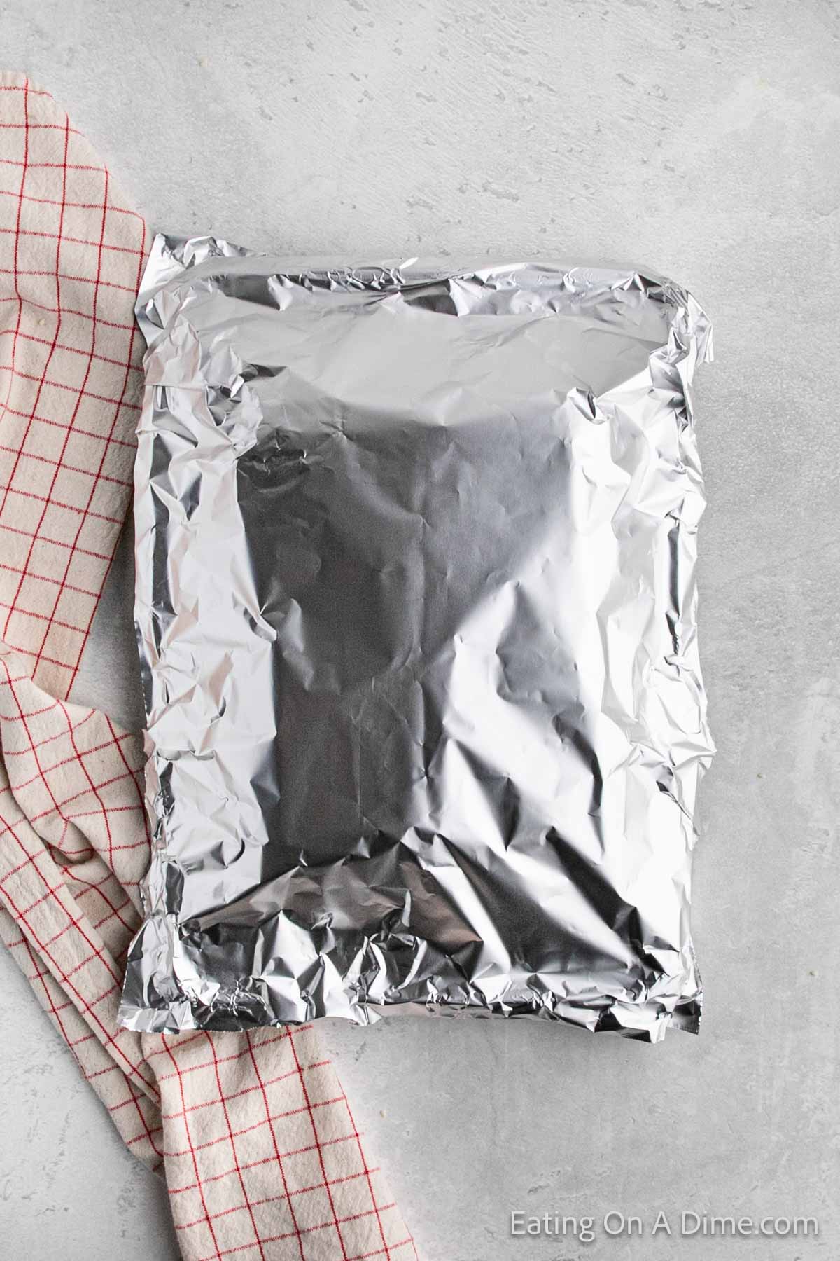 A sealed aluminum foil packet rests on a light gray surface, with a cream and red checkered cloth partially tucked under the left side of the packet. The foil is crinkled and shiny, reflecting light, hiding within it a delightful Hawaiian Roll Hamburger recipe waiting to be discovered.