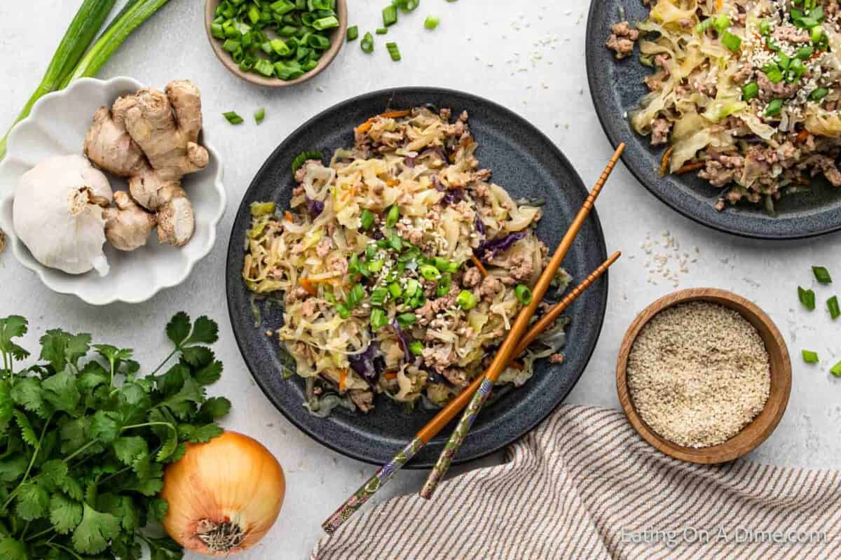 A top view of a meal featuring two plates filled with a stir-fry dish made with shredded vegetables, ground meat, and garnished with green onions and sesame seeds. Chopsticks rest on one plate. Surrounding ingredients include ginger, garlic, onions, cilantro, and a bowl of sesame seeds reminiscent of an instant pot southwest egg roll in a bowl.