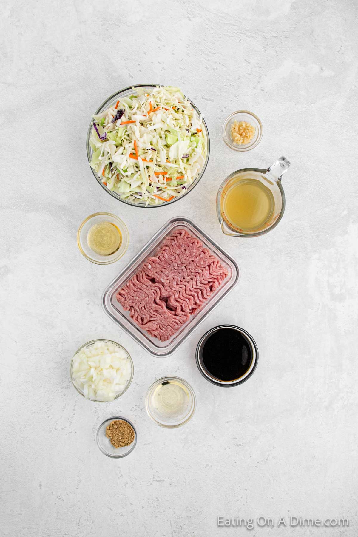 Top-down view of ingredients for an instant pot southwest egg roll in a bowl, including shredded cabbage and carrots, a jug of broth, minced garlic, sliced onions, ground meat, soy sauce, oil, spices, and salt. Everything is arranged on a light-colored surface.