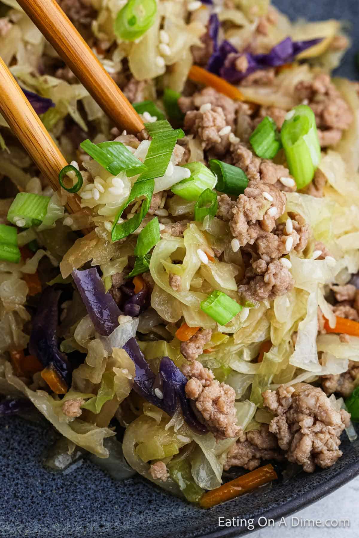 Close-up of an Instant Pot Southwest Egg Roll in a Bowl dish featuring ground meat, shredded cabbage, and carrots. The dish is garnished with chopped green onions and sesame seeds. Chopsticks are picking up a portion, showcasing the vibrant colors and textures of the ingredients.