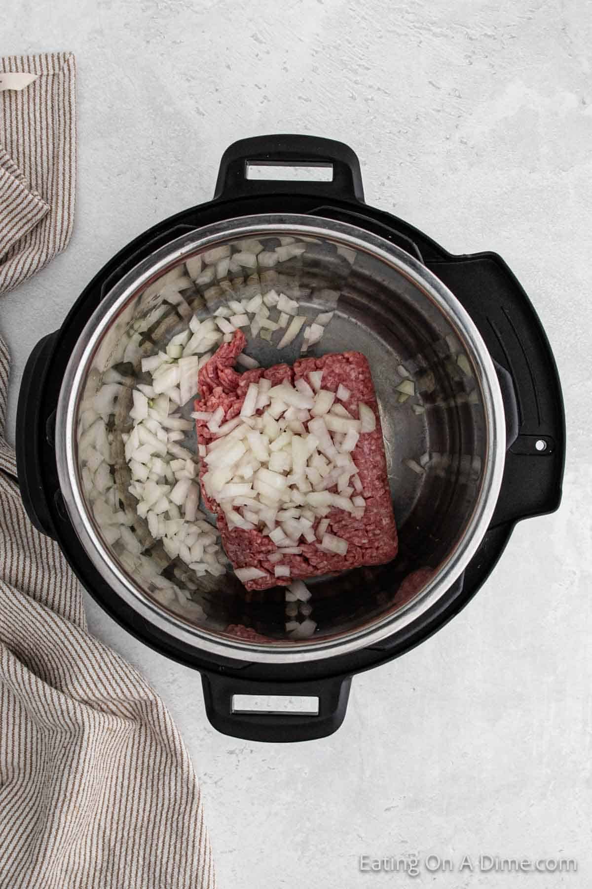 An overhead view shows an Instant Pot containing a block of ground meat with chopped onions on top, ready to become a savory Southwest Egg Roll in a Bowl. The pot is resting on a light-colored countertop, with a folded striped kitchen towel placed to the side.