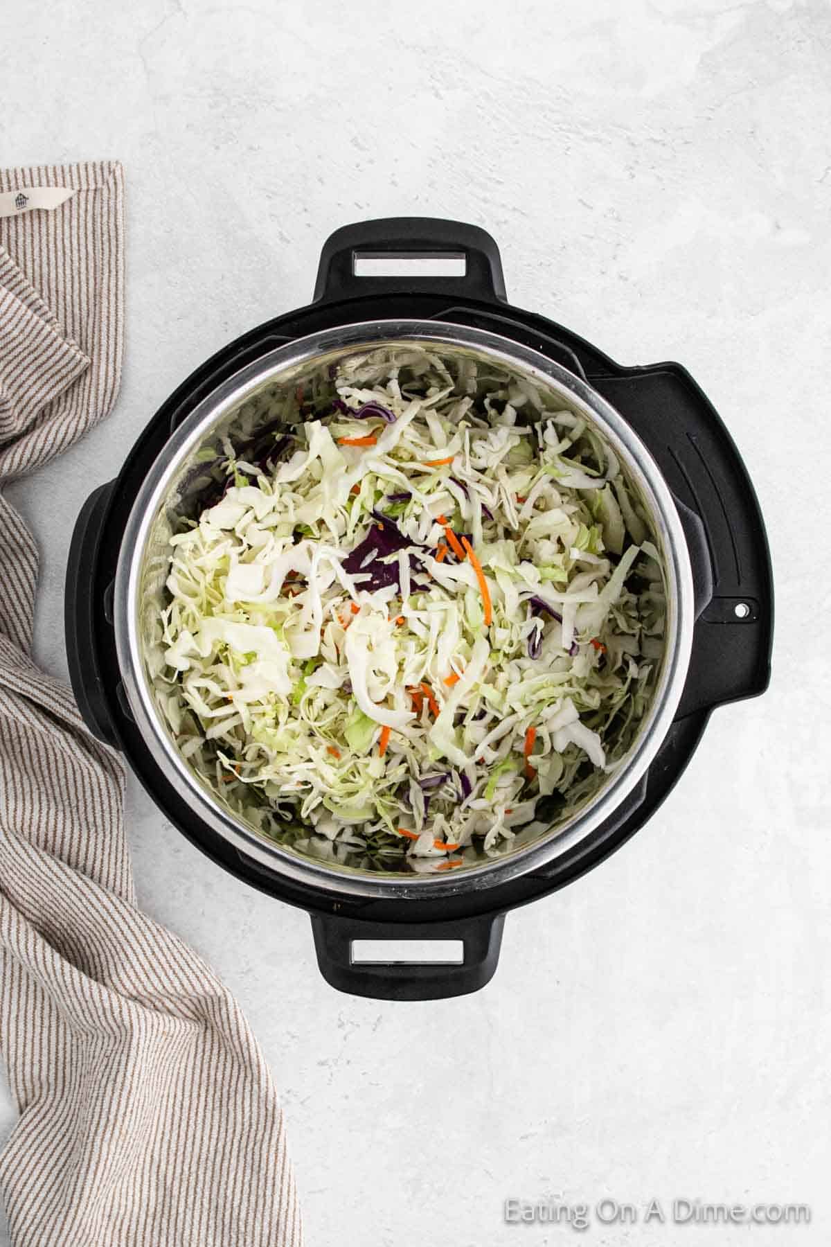 A black slow cooker containing a mix of shredded cabbage, carrots, and other vegetables sits on a light gray surface, evoking the vibrant flavors of an Instant Pot southwest egg roll in a bowl. A beige and white striped kitchen towel is placed to the left of the slow cooker.