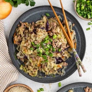 A dark gray plate filled with a cabbage stir-fry, reminiscent of an instant pot southwest egg roll in a bowl, featuring ground meat, chopped vegetables, and garnished with green onions and sesame seeds. Wooden chopsticks rest on the edge of the plate. Around the plate are sesame seeds, chopped green onions, an onion, and a kitchen towel.