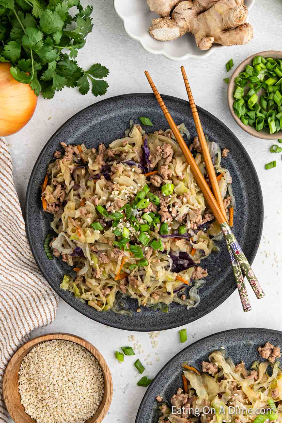 A top view of a plate filled with an Instant Pot Southwest egg roll in a bowl made of ground meat, shredded cabbage, and chopped vegetables. Two chopsticks rest on the plate's edge. Nearby are bowls of sesame seeds, chopped green onions, fresh ginger, and parsley.