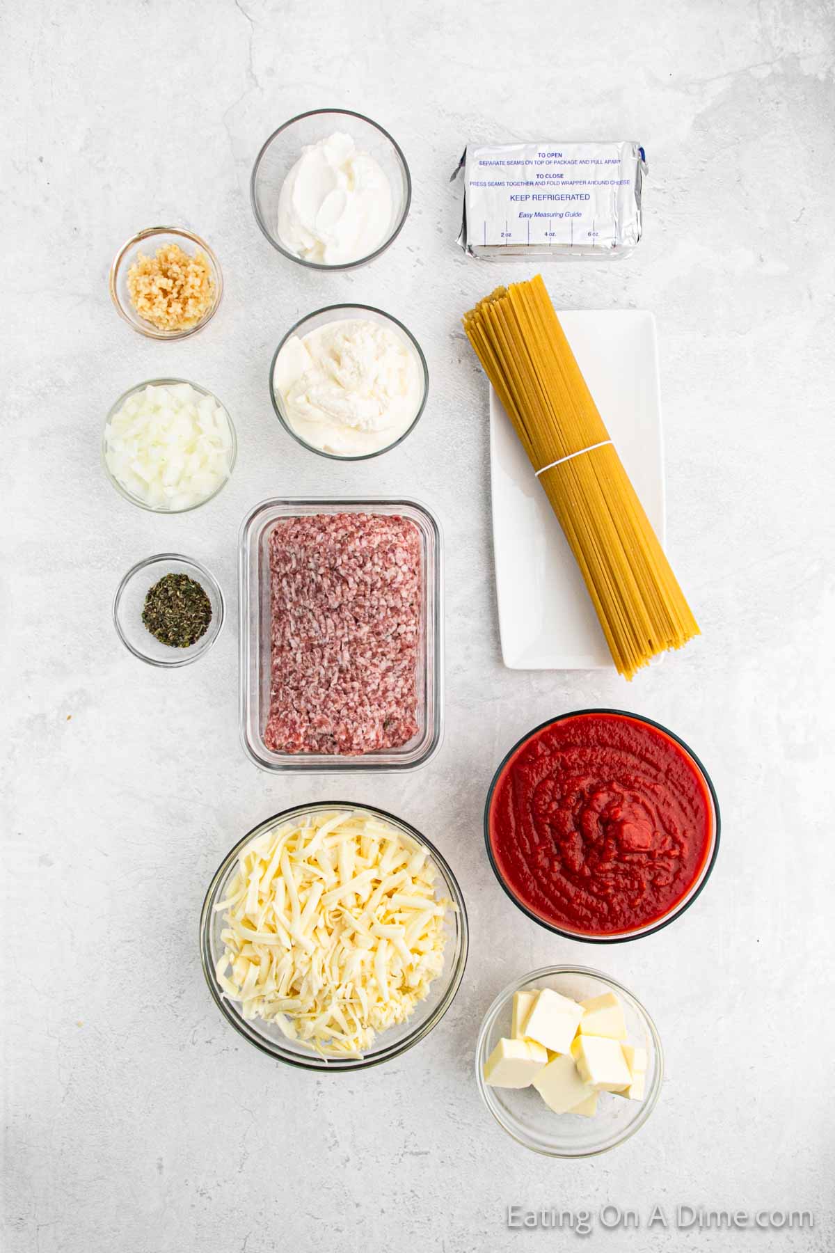 An arrangement of ingredients for a million dollar spaghetti recipe on a white countertop. Items include ground meat, shredded cheese, tomato sauce, spaghetti, butter, minced garlic, sour cream, ricotta cheese, chopped onions, and dried herbs.