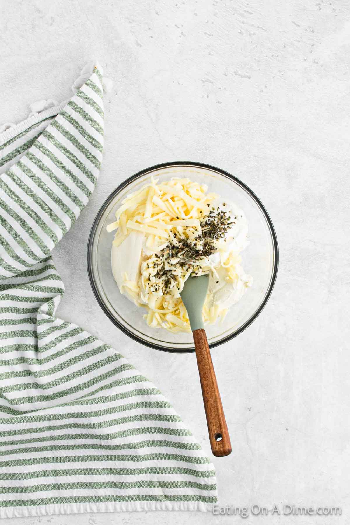 A glass bowl filled with shredded cheese, mayonnaise, and seasoning is being mixed with a green spatula. The bowl is placed on a light-colored surface next to a green and white striped cloth, reminiscent of the million dollar spaghetti recipe's rich ingredients.