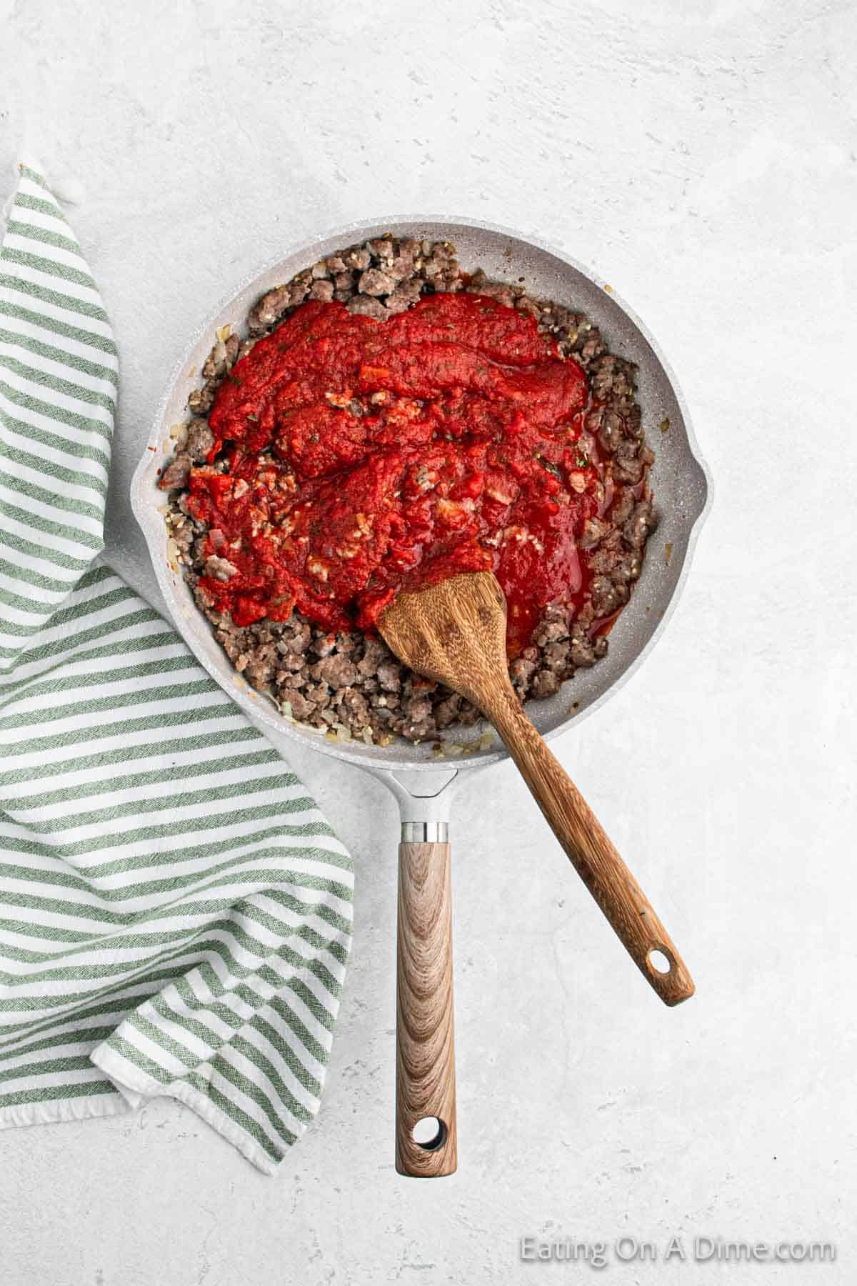 A skillet filled with cooked ground meat topped with tomato sauce, resembling a hearty million dollar spaghetti recipe, with a wooden spoon resting inside the skillet. Next to the skillet is a green and white striped towel, all set on a light-colored countertop.