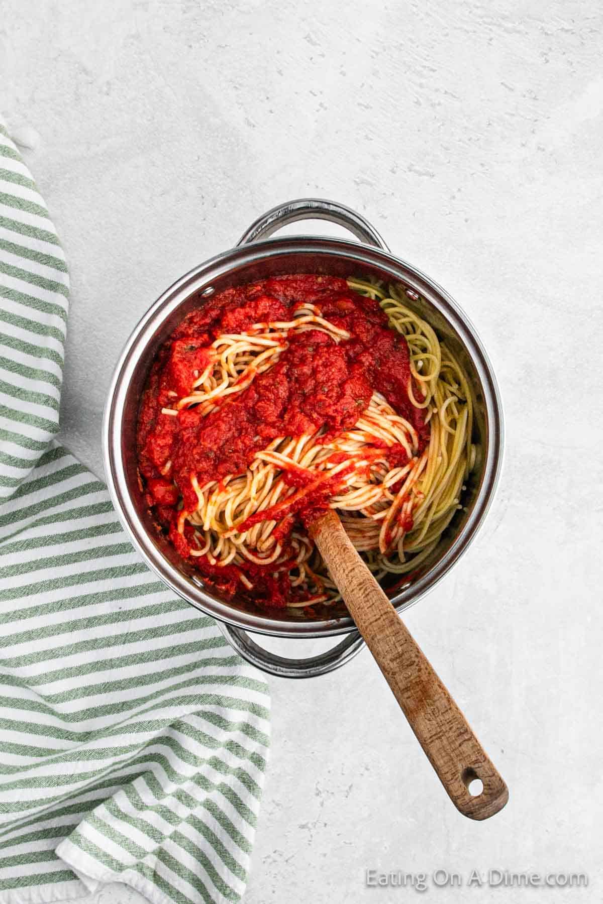 A stainless steel pot filled with cooked spaghetti intertwined with rich marinara sauce sits on a light gray surface. A wooden spoon rests in the pot, hinting at the secret to this million dollar spaghetti recipe. A green and white striped cloth is partially visible to the side.
