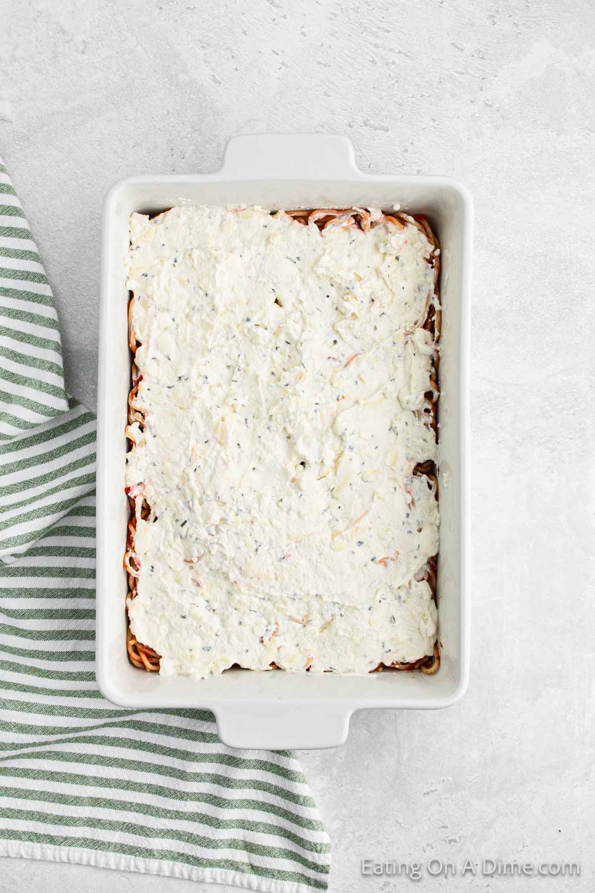 A rectangular white baking dish filled with a creamy, white mixture on top of layered ingredients. This million dollar spaghetti recipe is placed on a light gray surface next to a green and white striped fabric. The edges of the baking dish have handles.