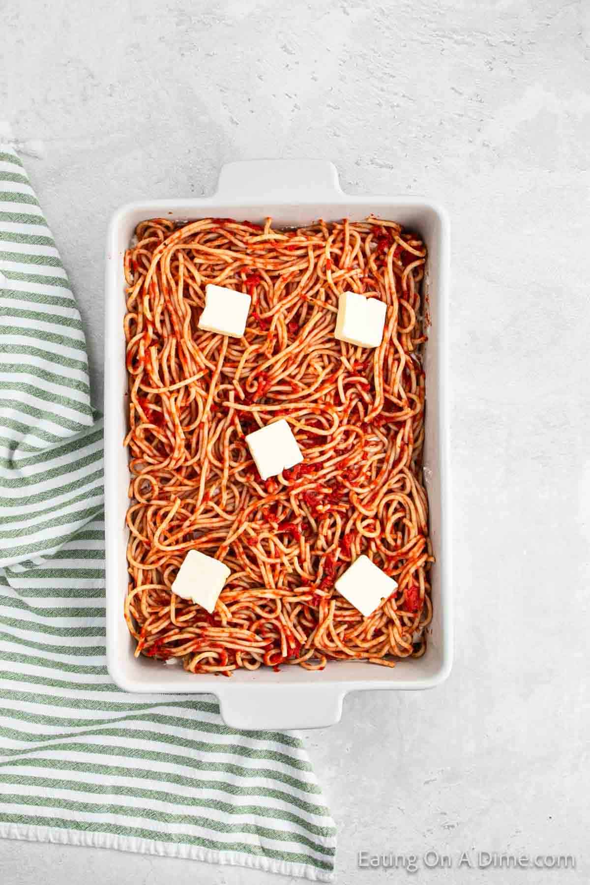 A rectangular white baking dish filled with cooked spaghetti and topped with six small squares of butter, showcasing a million dollar spaghetti recipe. The dish is placed on a light gray surface next to a folded, striped green and white kitchen towel.