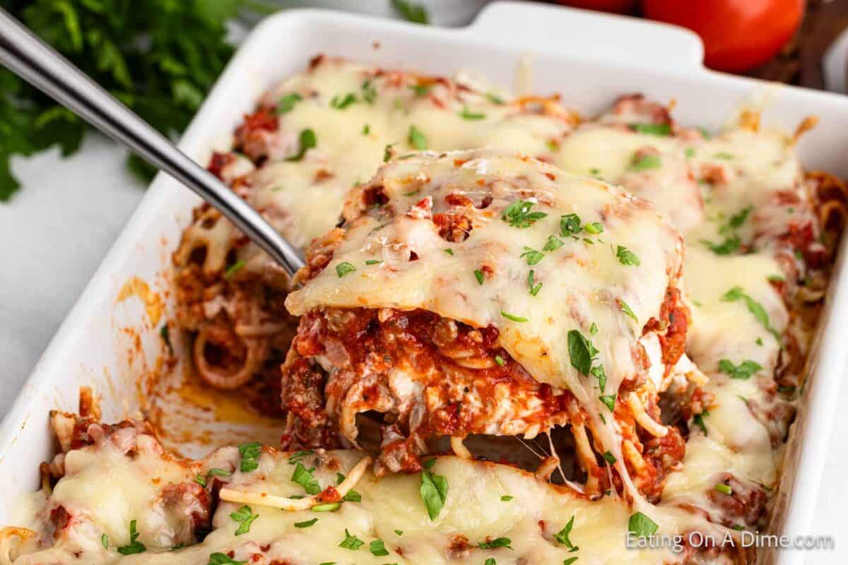 A close-up of a baked spaghetti dish in a rectangular white dish. A serving is being lifted out with a metal spatula, showcasing layers of spaghetti, meat sauce, and melted cheese sprinkled with chopped parsley. Fresh parsley and tomatoes are in the background. It's the perfect million dollar spaghetti recipe.