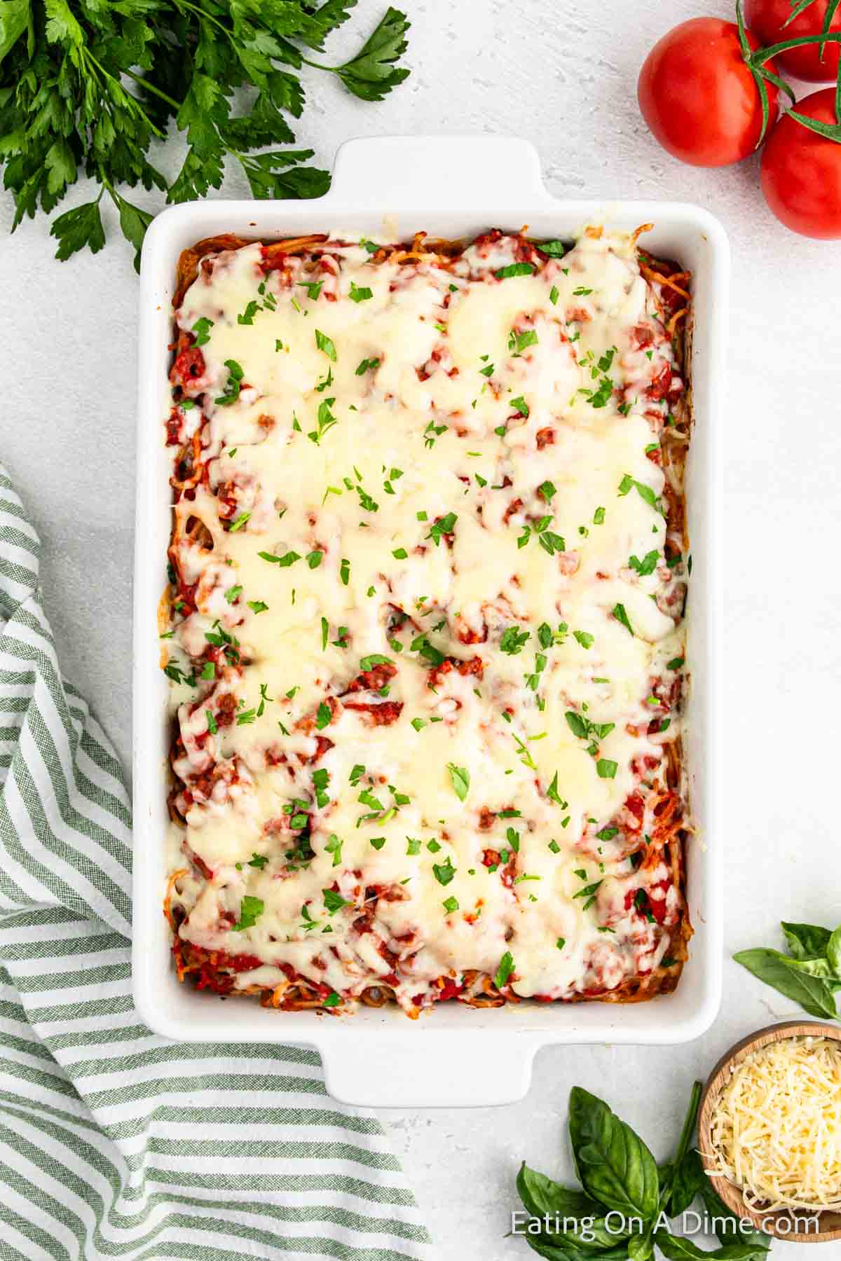 A white rectangular baking dish filled with a freshly baked million dollar spaghetti casserole topped with melted cheese and chopped herbs. Surrounding the dish are sprigs of parsley, whole tomatoes, a dish of shredded cheese, and a green striped kitchen towel.