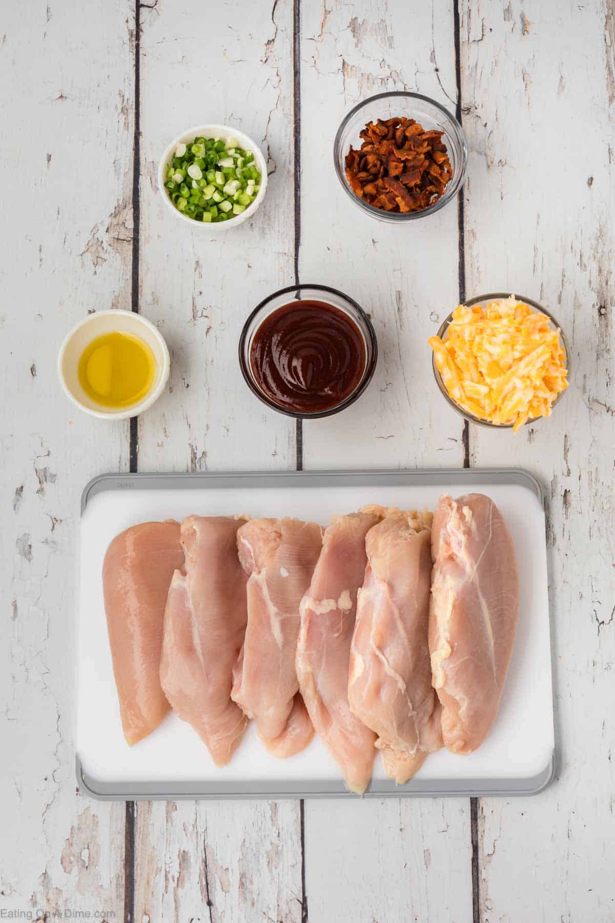 Ingredients - Raw chicken breast on a cutting board, bowl of shredded cheese, BBQ Sauce, oil, chopped green onions and chopped bacon
