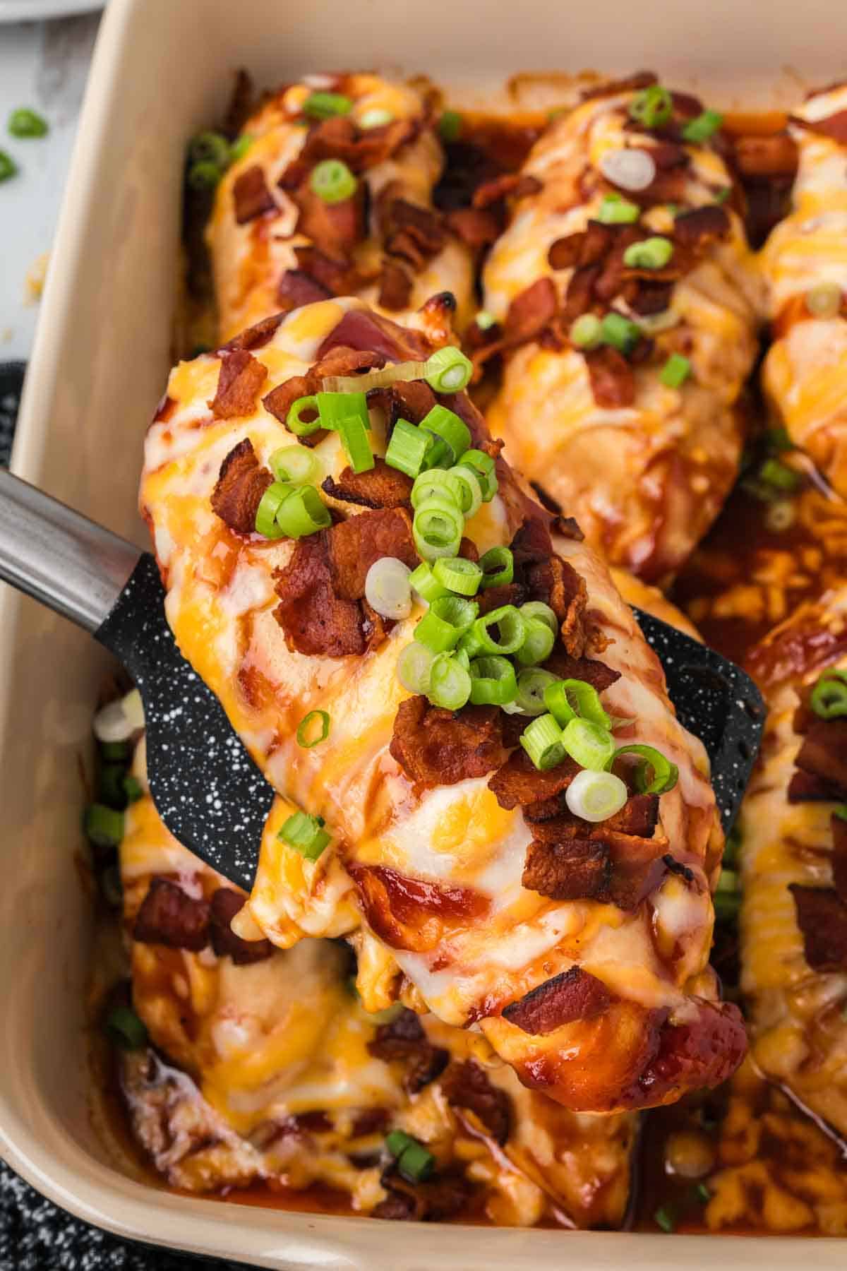 Baked chicken topped with chopped bacon, green onions, BBQ Sauce and melted cheese in a baking dish with a serving on a spatula