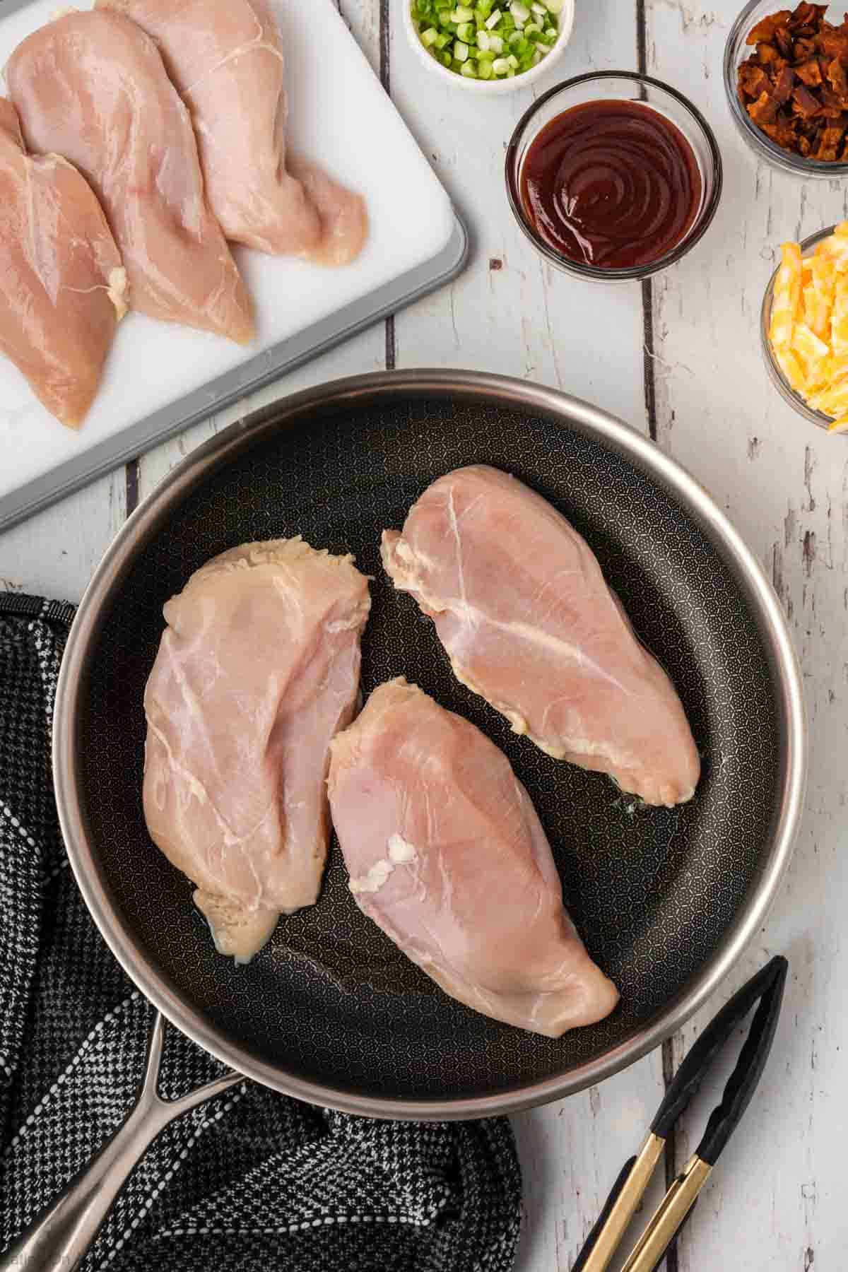 Cooking chicken breast in a skillet with a cutting board of chicken breast and bowls of BBQ sauce, chopped bacon, shredded cheese and green onions