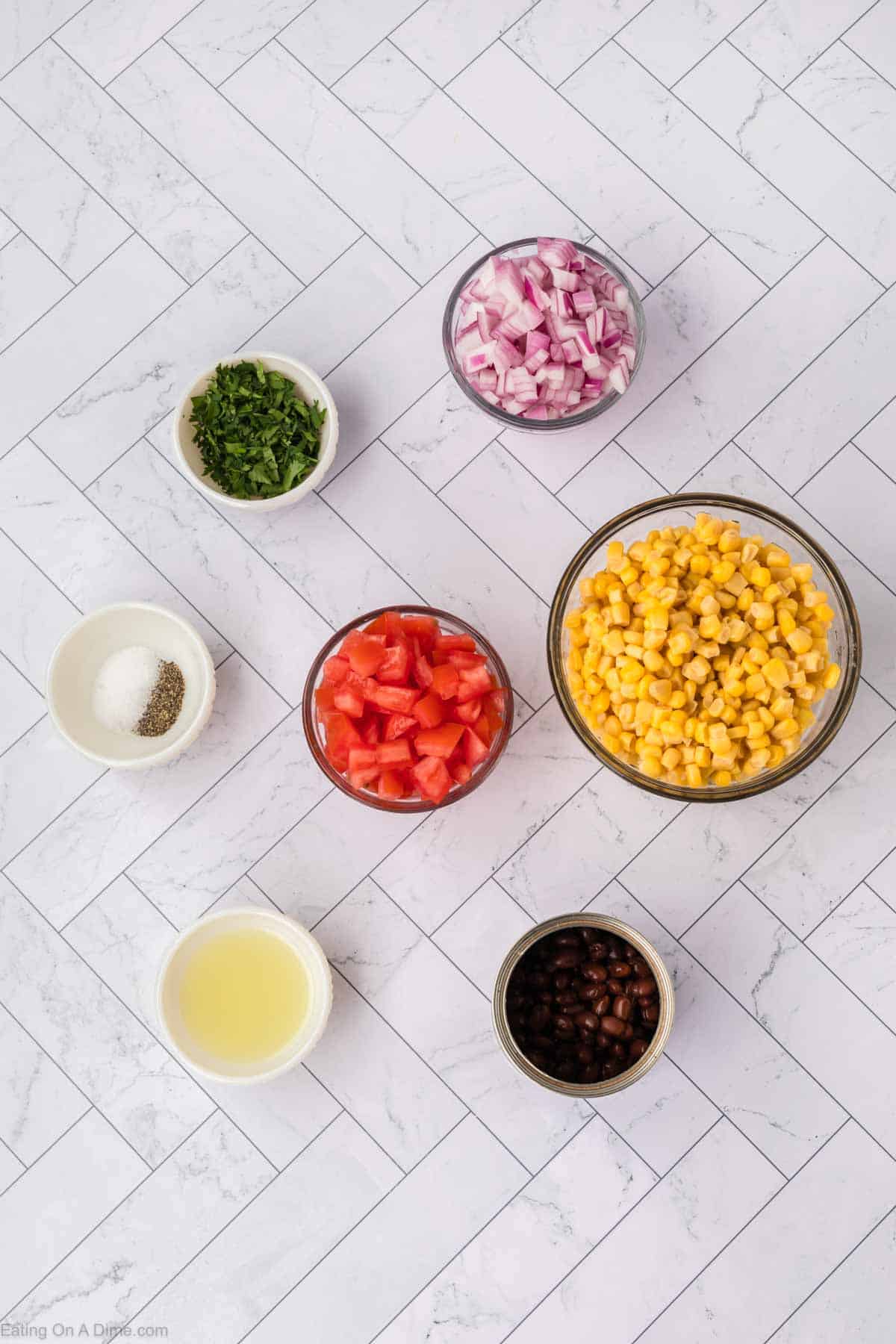 Ingredients for a delicious Black Bean Salsa are laid out on a white tiled surface. They include chopped red onions, corn, diced tomatoes, black beans, chopped cilantro in small bowls, ground black pepper and salt in a small dish, and lemon juice in a cup.