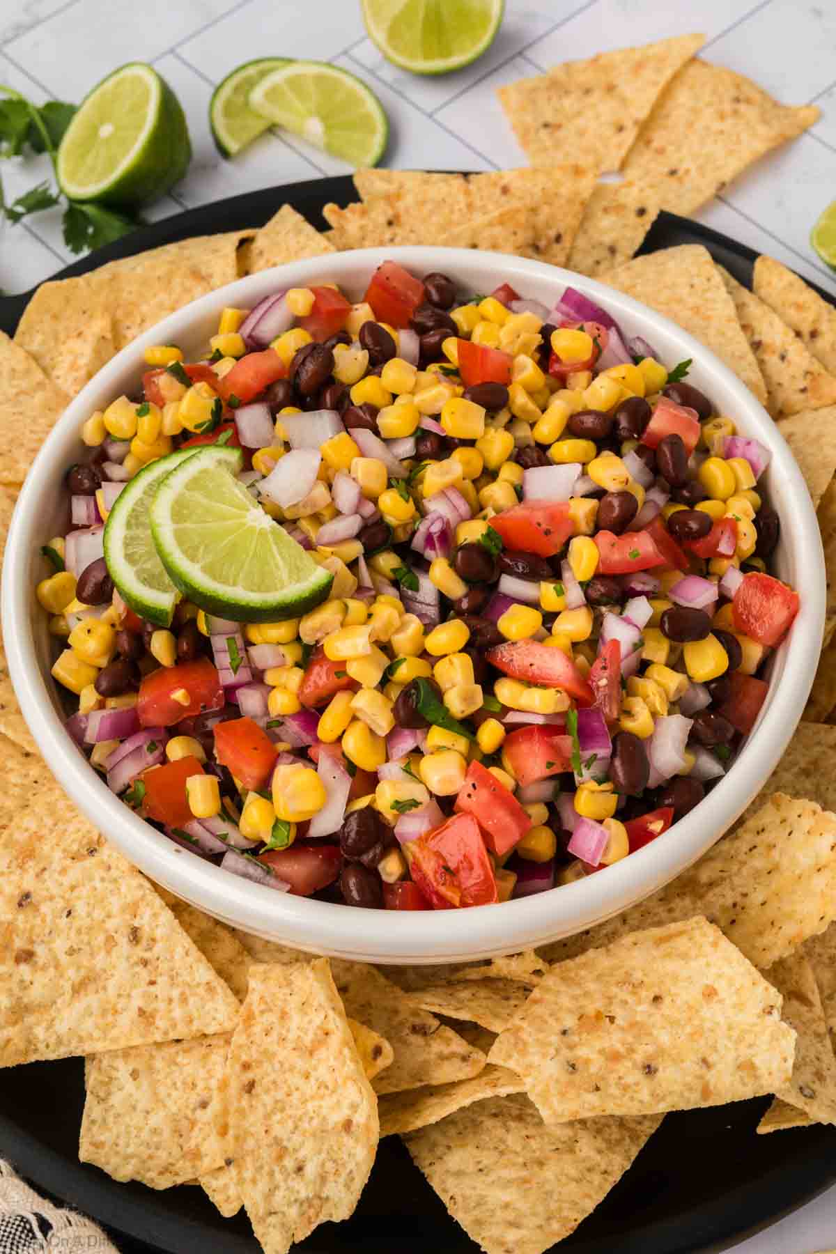 A bowl of colorful black bean salsa with corn, onions, tomatoes, and chopped cilantro, garnished with lime slices. The bowl is surrounded by tortilla chips, with lime halves in the background on a light countertop.