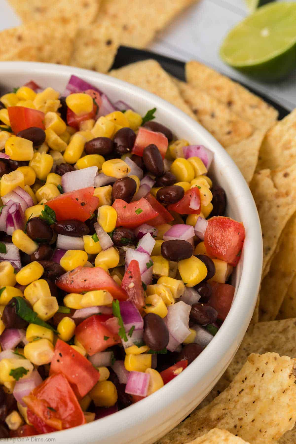 A close-up of a bowl filled with vibrant black bean salsa, featuring corn, black beans, diced tomatoes, red onions, and cilantro. The bowl is surrounded by tortilla chips, and there are lime wedges in the background.