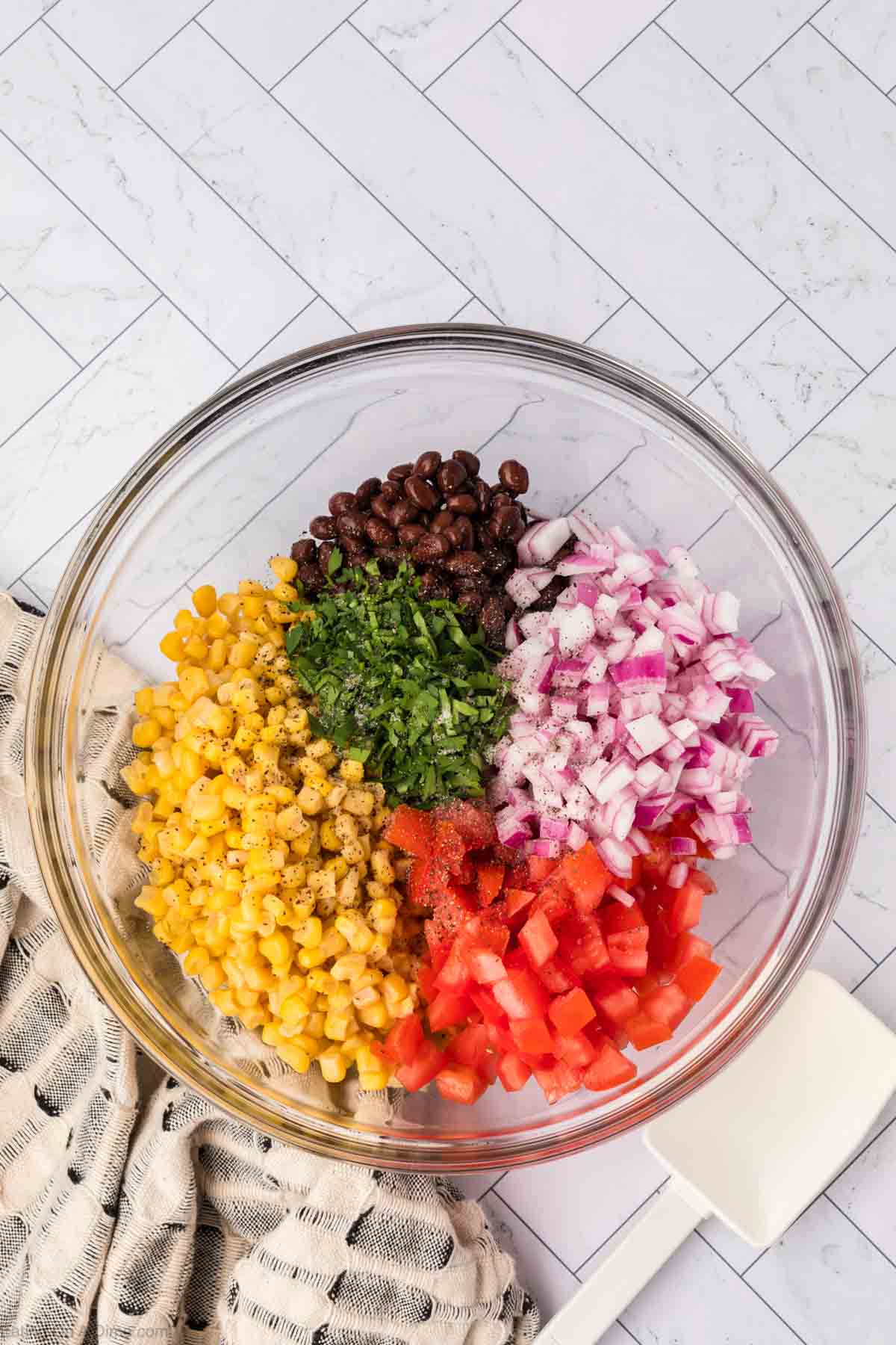 A glass bowl contains prepped ingredients for a delicious black bean salsa: black beans, diced red onion, chopped tomatoes, corn kernels, and fresh cilantro. The bowl is placed on a patterned tiled surface next to a white spatula and a textured cloth.
