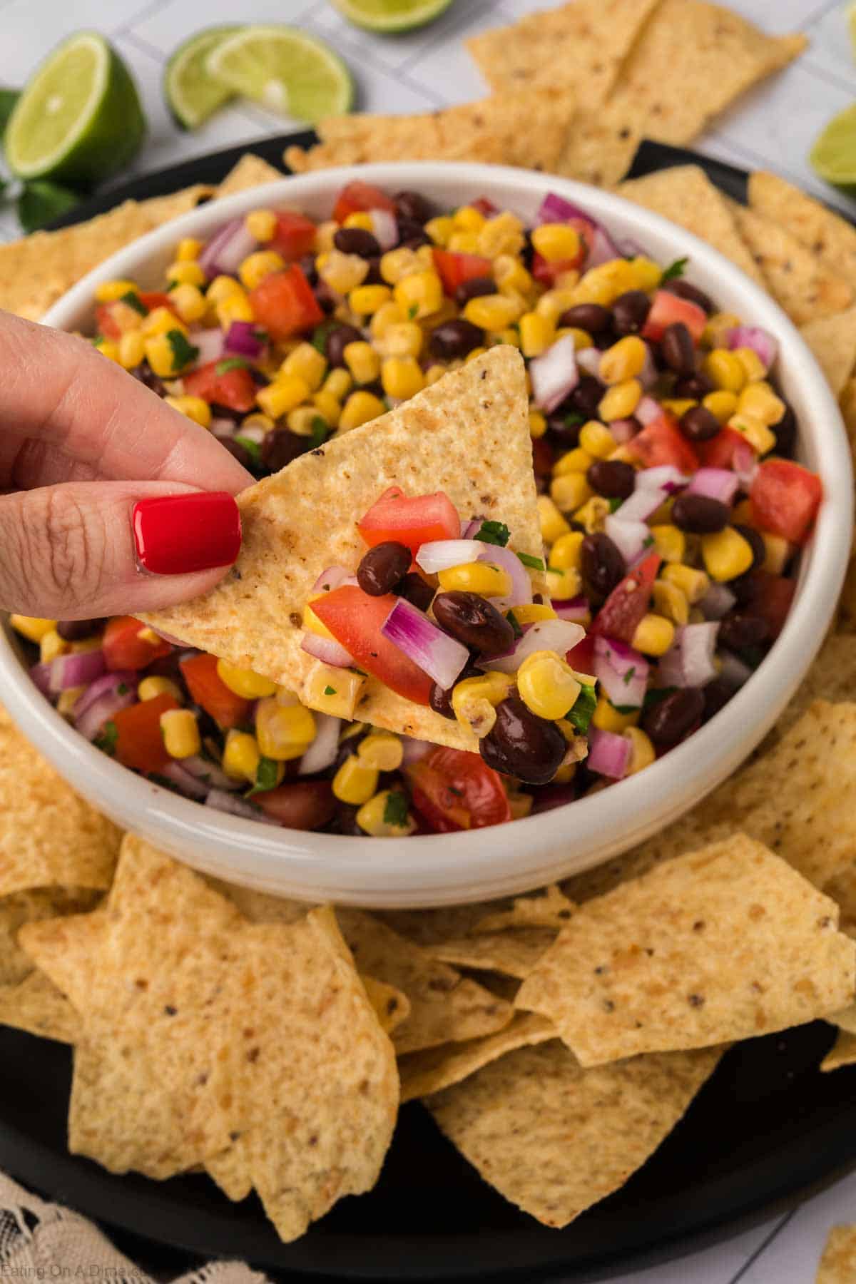 A hand with red nail polish holds a tortilla chip topped with vibrant black bean salsa, featuring a colorful mix of corn, tomatoes, onions, and cilantro. The chip is lifted from a white bowl filled with the same mixture, surrounded by additional tortilla chips and lime wedges.