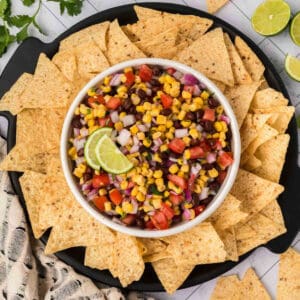 A bowl of vibrant black bean salsa, including corn, black beans, tomatoes, onions, and cilantro, garnished with two lime slices. The bowl is surrounded by a ring of tortilla chips on a round black plate. Lime halves and cilantro leaves are artfully placed in the background.