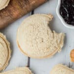 A round, crustless sandwich with a bite taken out of it, placed on a white wooden surface. To the right is a small dish of dark purple jam, and part of a wooden rolling pin is visible in the top-left corner. Other homemade uncrustables are partially visible at the edges.