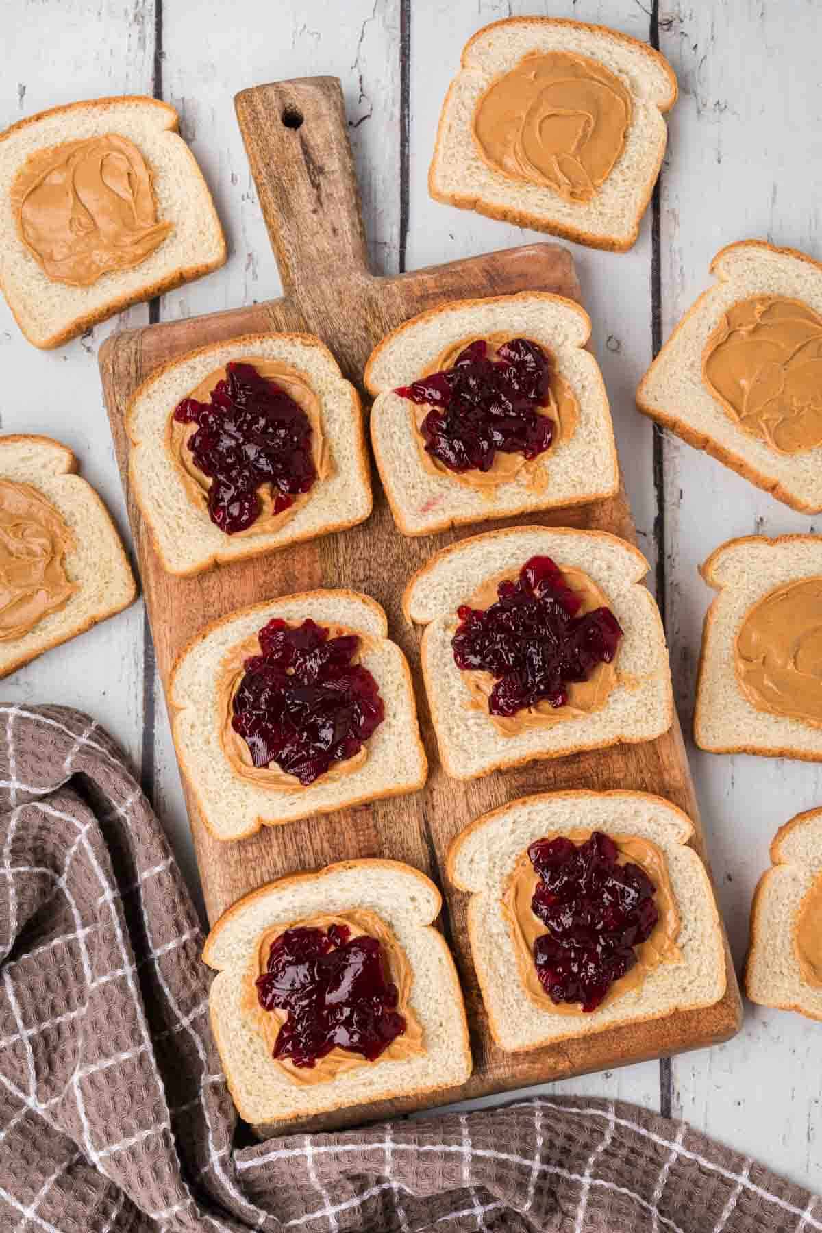 Slices of bread are placed on a wooden cutting board and surrounding surface, each topped with either peanut butter or a combination of peanut butter and grape jelly, resembling homemade uncrustables. A light brown checkered cloth lies next to the cutting board.