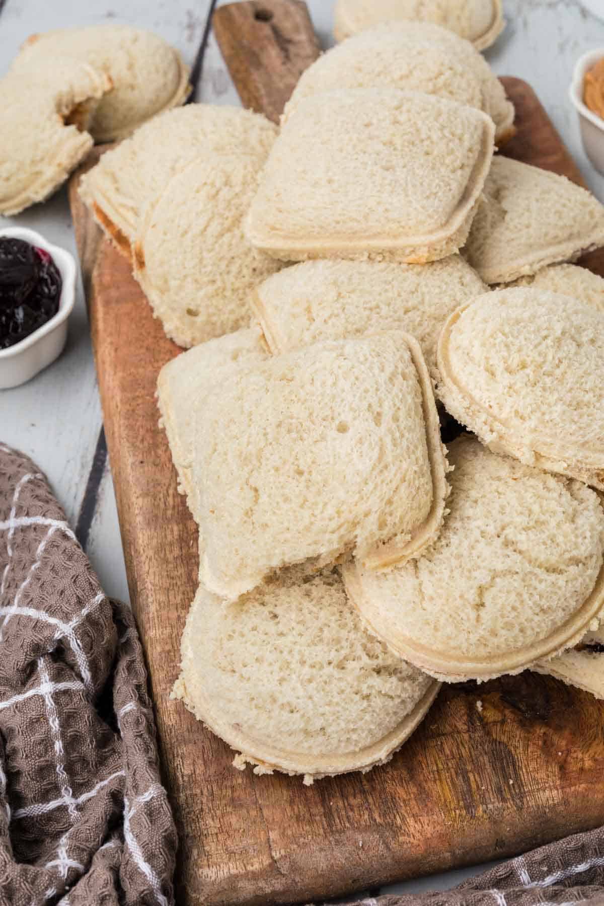 A wooden board is piled with several pieces of crustless, square, and round sandwich bread. A bowl of dark jam and a partially visible bowl of peanut butter suggest the beginning stages of homemade uncrustables. A brown hand towel with a plaid pattern is draped on the left side of the board.
