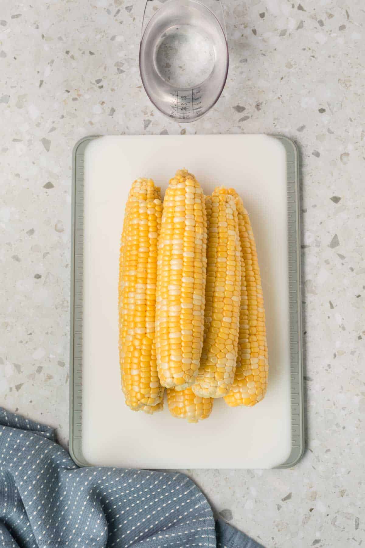 Corn on the cob stacked on a platter with a measuring cup of water