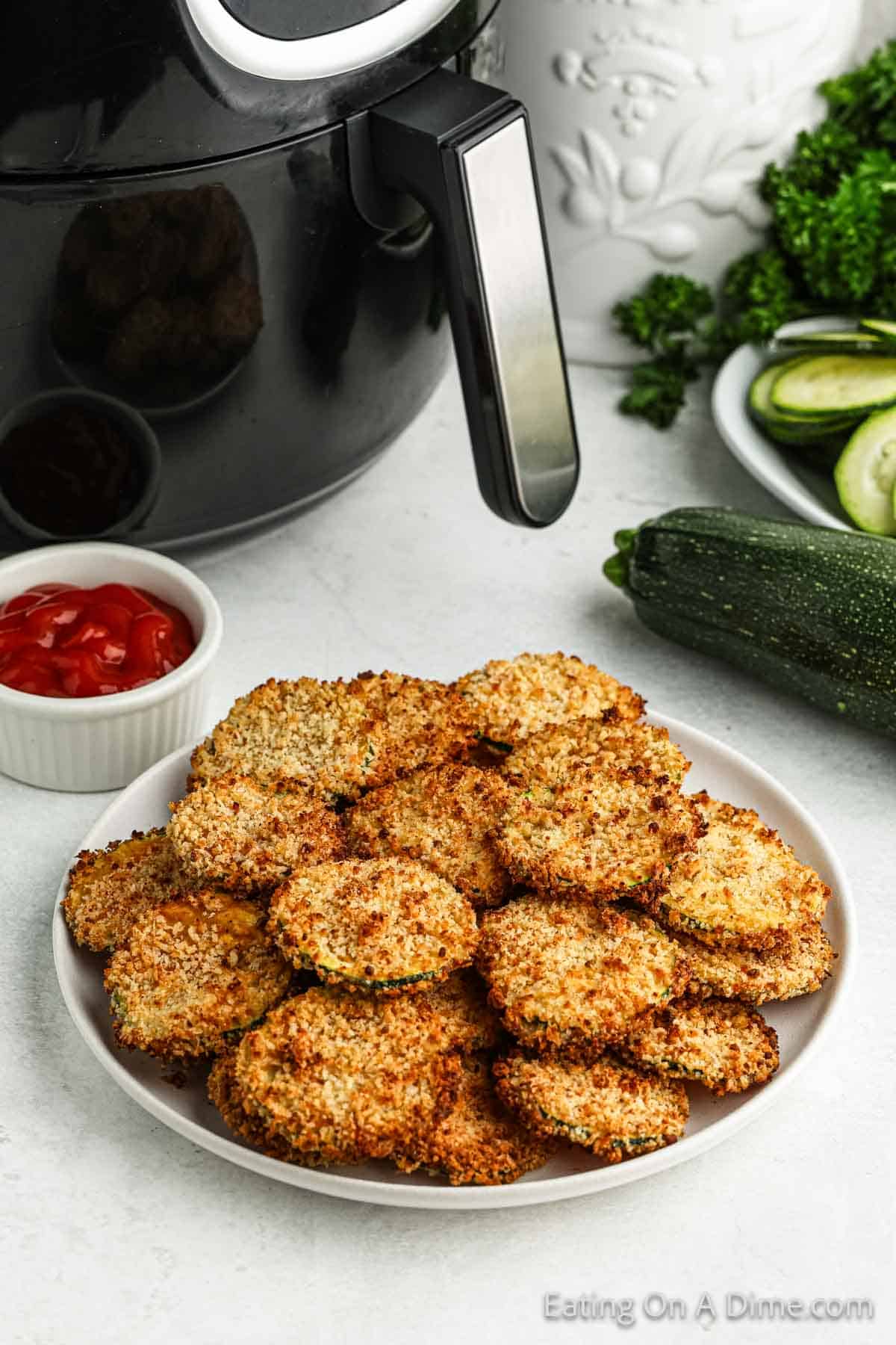 Crispy Zucchini Chips stacked on a plate with a small of bowl of ketchup and the air fryer in the background