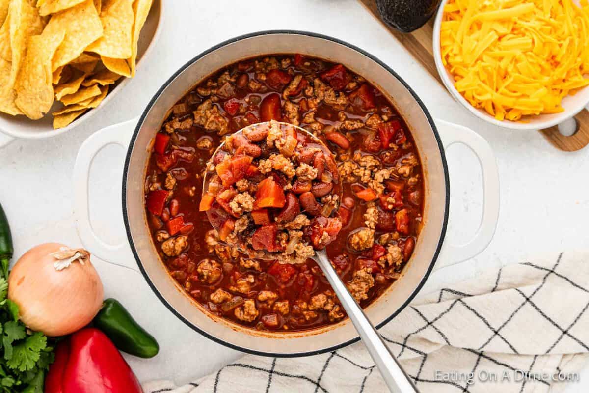 A large pot of ground turkey chili filled with tomatoes and beans. A ladle scoops up a portion. Surrounding the pot are bowls of shredded cheese and tortilla chips, and fresh vegetables like onion and peppers are nearby.