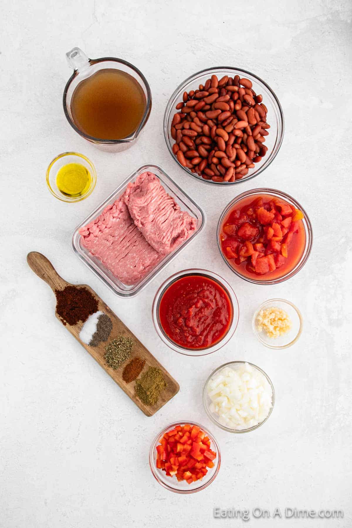 Top view of ground turkey chili ingredients, including ground meat, kidney beans, diced tomatoes, tomato sauce, chopped onions, minced garlic, red bell peppers, various spices, olive oil, and broth arranged on a light surface.