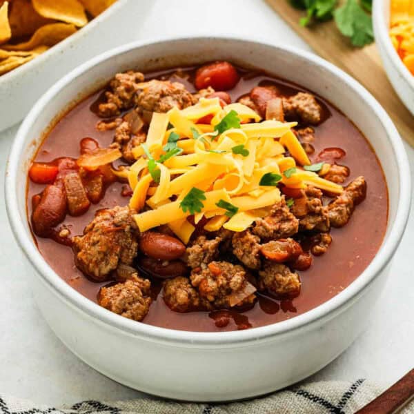 A bowl of ground turkey chili topped with shredded cheddar cheese and garnished with parsley. The hearty chili includes kidney beans and tomatoes, offering a delicious twist. A side of tortilla chips is partially visible in the background.