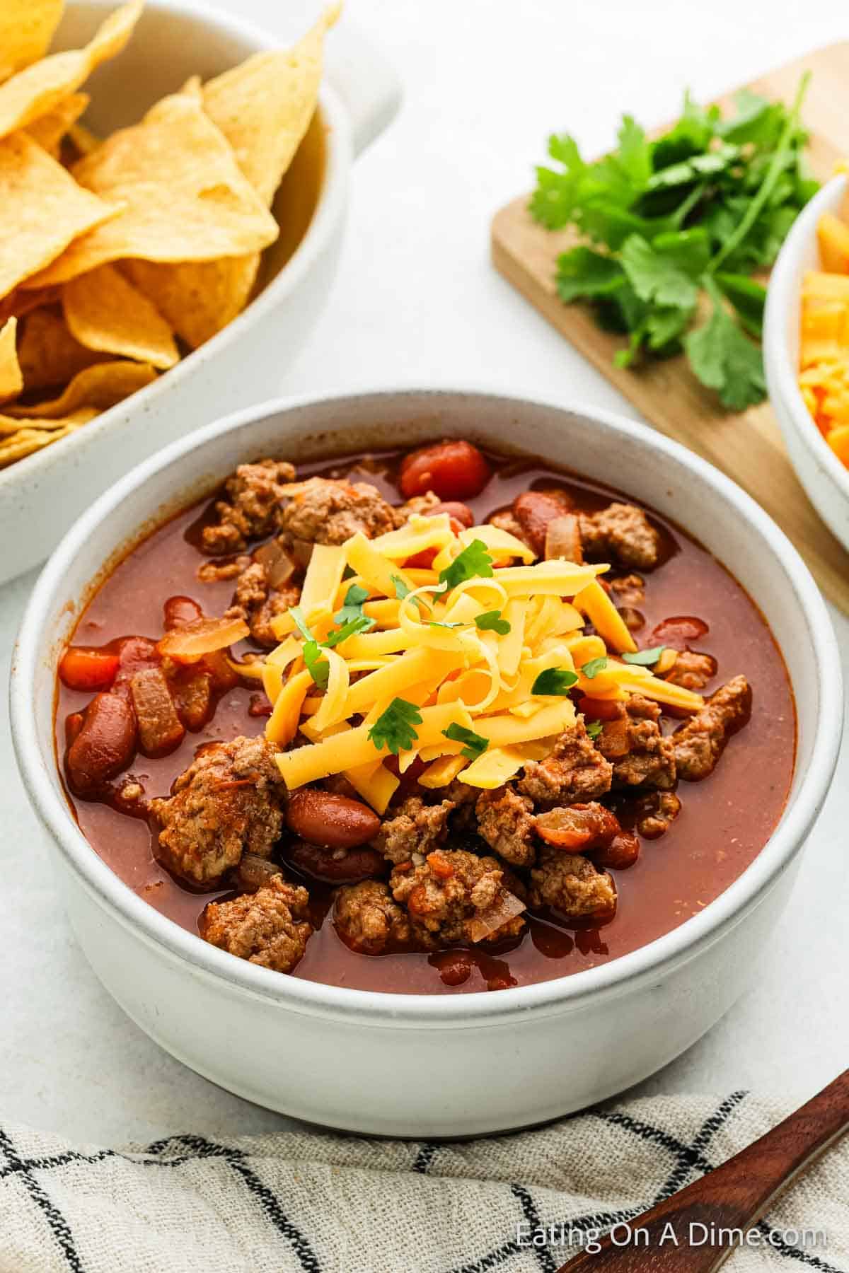 A bowl of ground turkey chili topped with shredded cheese and cilantro. Surrounding the bowl are crispy tortilla chips, a small dish of shredded cheese, and a few sprigs of fresh cilantro on a wooden surface.