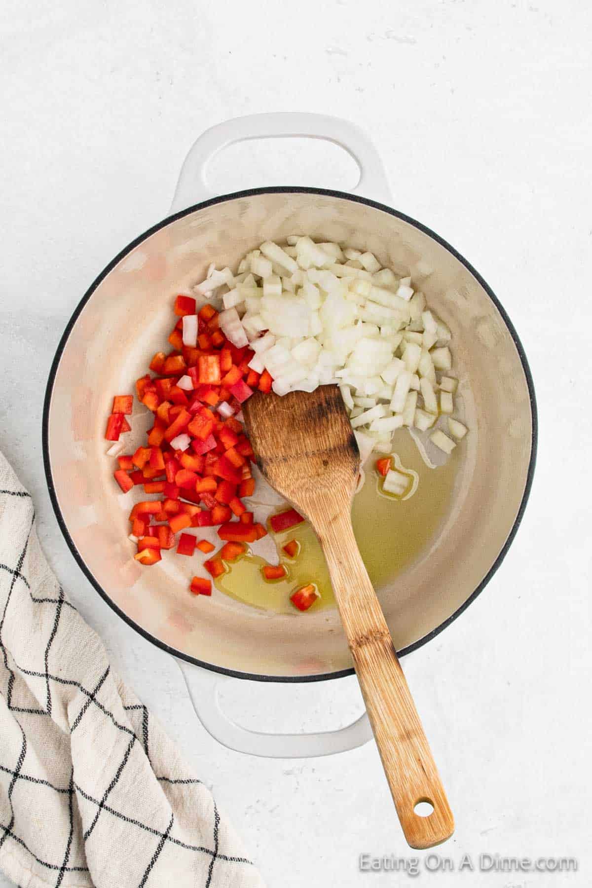 A white pot on a light surface contains chopped red bell peppers and onions, with a wooden spatula resting inside, ready to stir in the ground turkey chili. A striped kitchen towel is partially visible on the left.