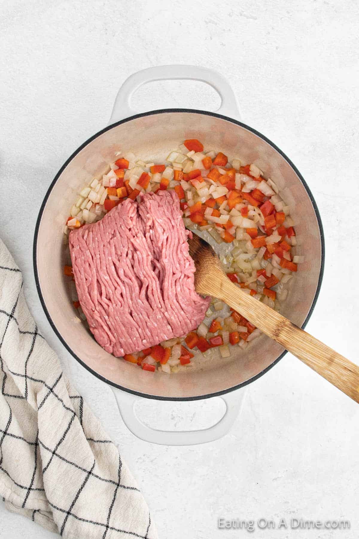 A large white pot contains chopped onions and red bell peppers with a block of ground turkey chili on top. A wooden spoon rests in the pot. A white dish towel with black lines is nearby on a light-colored surface.