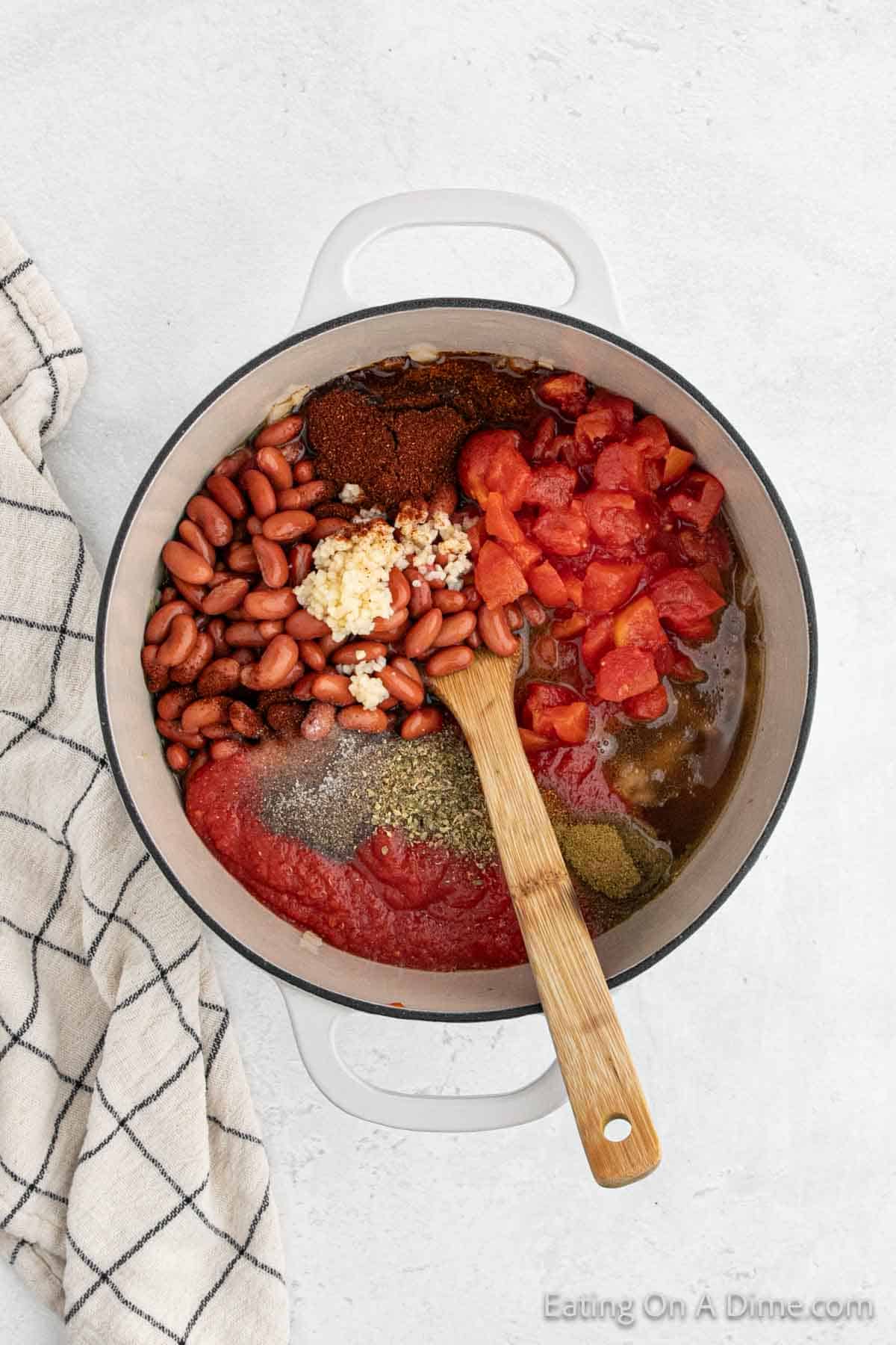 A white pot on a white surface holds ingredients for a delicious ground turkey chili, featuring kidney beans, chopped tomatoes, minced garlic, tomato paste, and herbs. A wooden spoon rests in the mix while a checkered kitchen towel lies nearby.