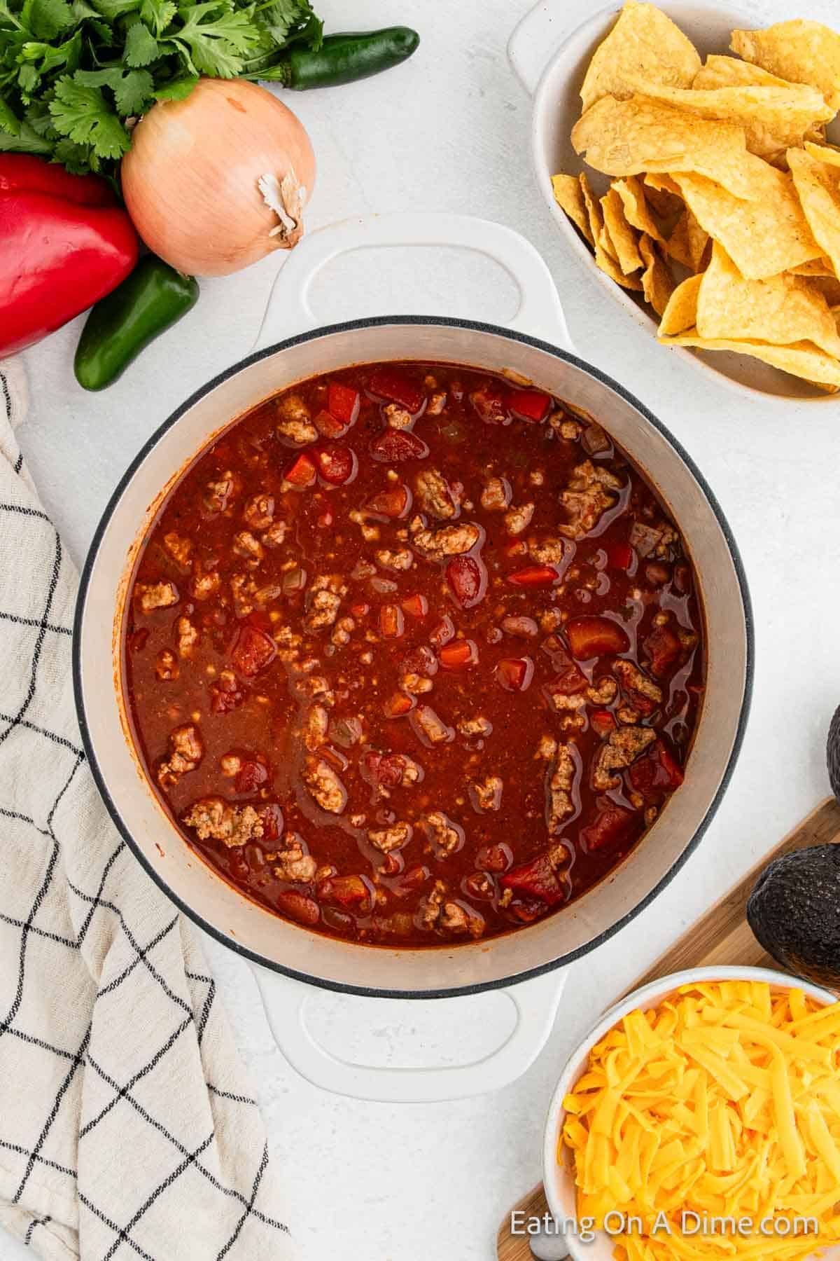 A pot of hearty ground turkey chili with meat and tomatoes sits on a white table. Surrounding it are tortilla chips in a bowl, shredded cheese, avocado, a red pepper, onion, jalapeños, cilantro, and a checkered dish towel.