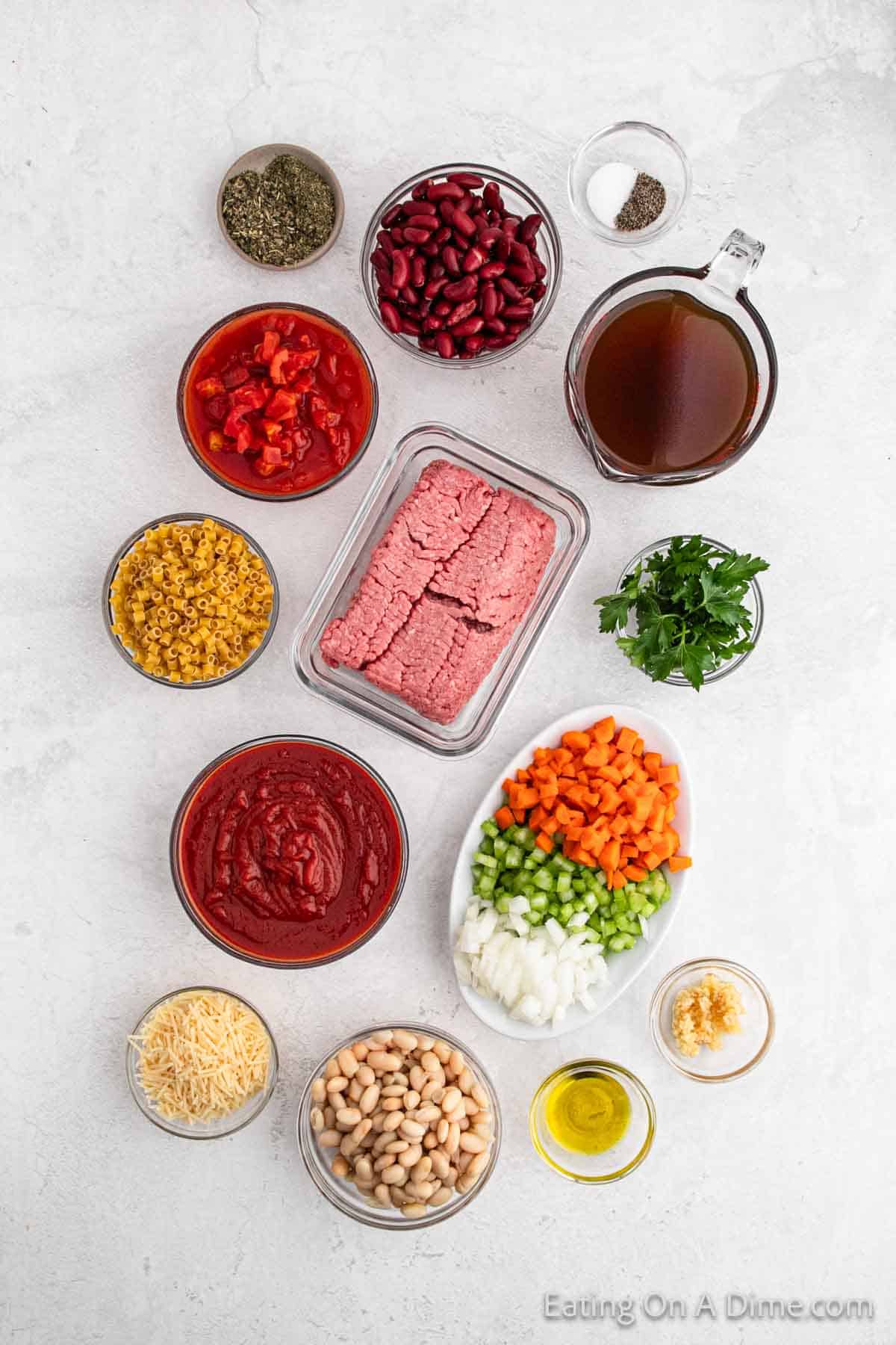 A flat lay of Pasta Fagioli Soup ingredients on a white surface, featuring ground meat, kidney beans, white beans, diced tomatoes, beef broth, macaroni, tomato sauce, diced carrots, diced celery, diced onions, shredded cheese, olive oil, parsley and seasonings.