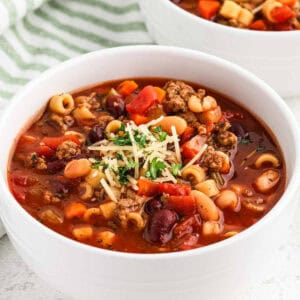 A white bowl filled with hearty minestrone soup, featuring ingredients like macaroni pasta, beans, ground meat, diced tomatoes, and a garnish of shredded cheese and chopped parsley. The Pasta Fagioli Soup sits invitingly as a second bowl is partially visible in the background.