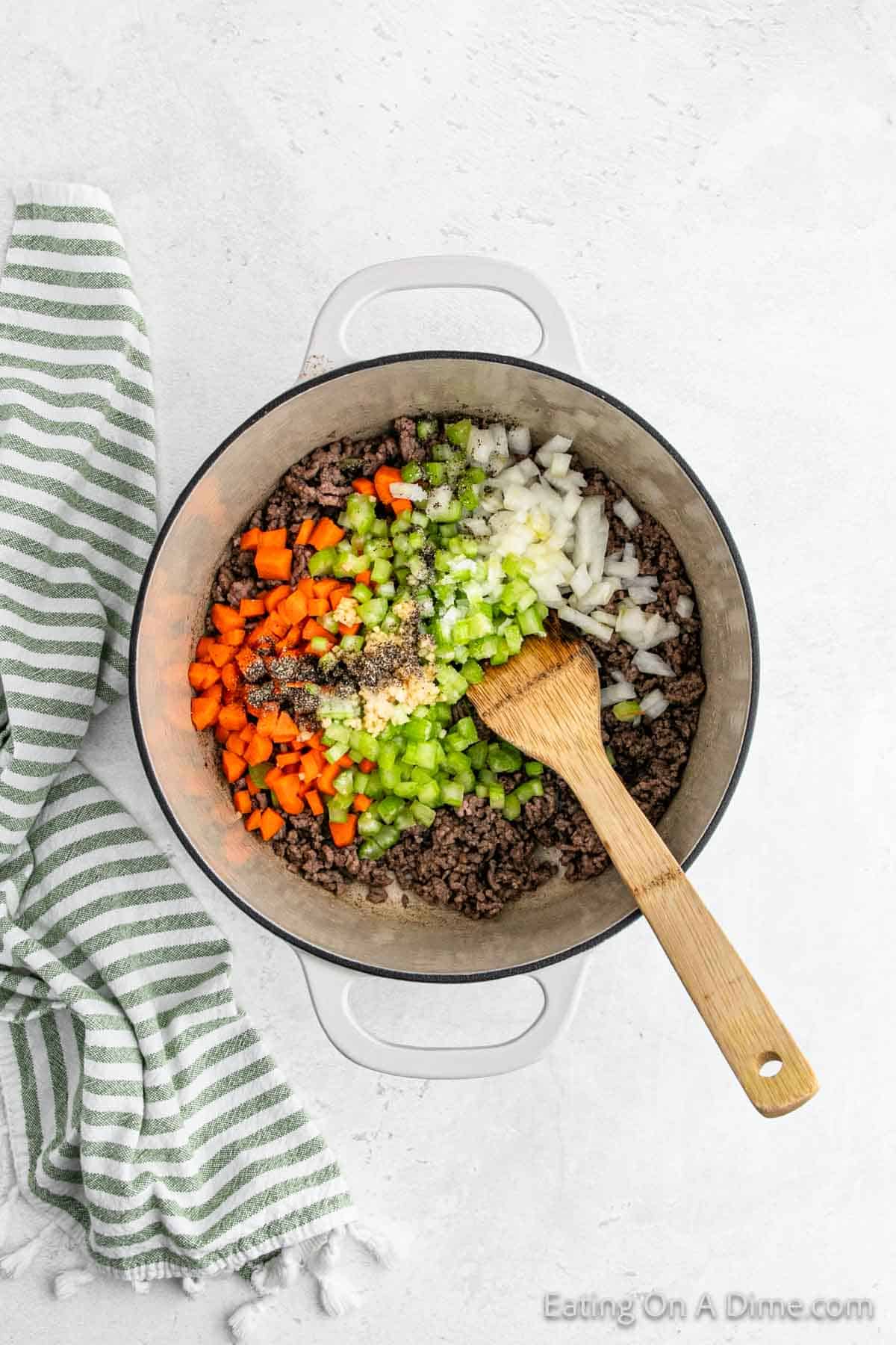 A pot on a light-colored surface containing browned ground beef, diced carrots, celery, and onions, with seasoning sprinkled on top—perfect for a hearty Pasta Fagioli Soup. A wooden spoon rests inside the pot. A green and white striped towel is placed beside it.