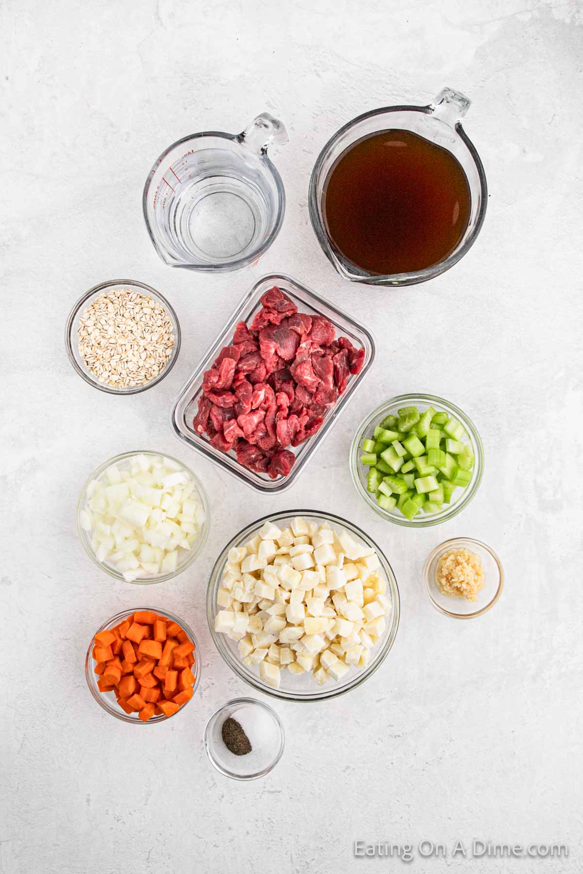 Ingredients for crockpot beef barley soup, including diced onions, chopped carrots, diced celery, minced garlic, cubed beef, diced potatoes, beef broth, water, uncooked barley, and pepper—arranged separately in clear containers on a light-colored surface.