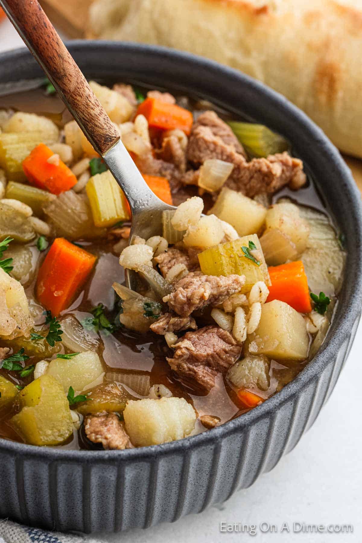 A close-up of a hearty crockpot beef barley soup served in a dark bowl. The soup is filled with chunks of beef, diced potatoes, carrots, celery, and barley, all in a rich broth. A spoon is dipping into the bowl, showcasing the ingredients. A piece of bread is in the background.