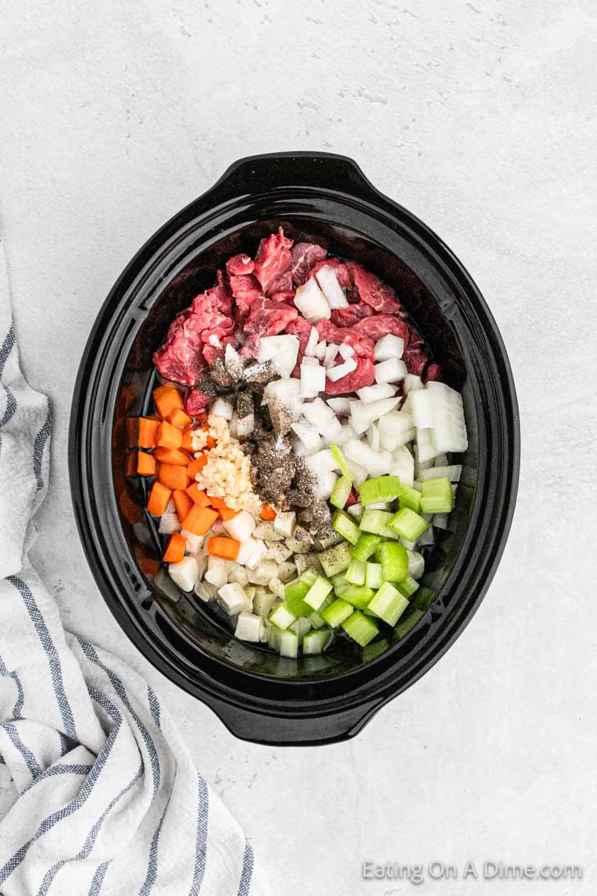 A black slow cooker is filled with chopped ingredients including raw beef, onions, carrots, celery, and minced garlic. Seasonings are sprinkled on top for a mouth-watering crockpot beef barley soup. The slow cooker is placed on a light, textured surface next to a striped kitchen towel. The text "Eating On A Dime" is visible in the bottom right corner of the image.