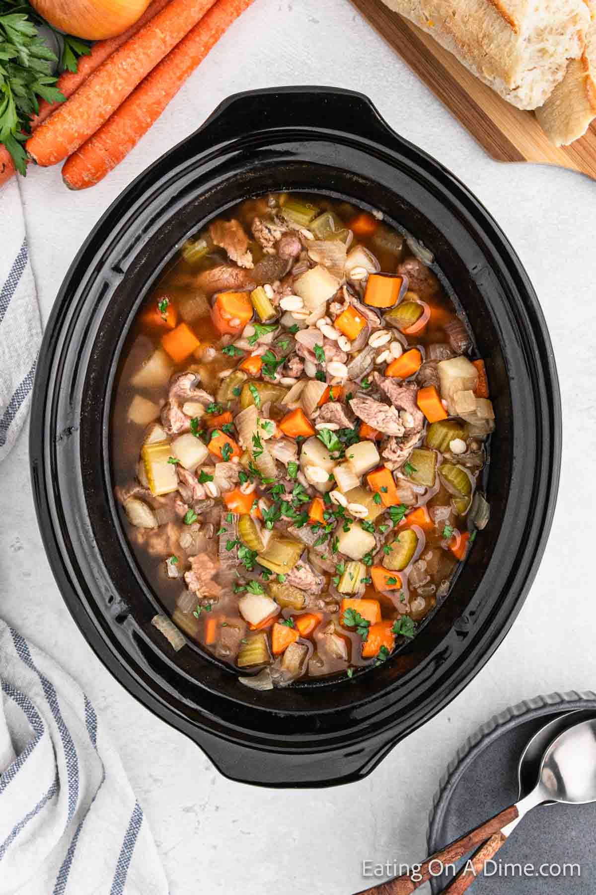 A crockpot filled with a hearty beef stew, featuring chunks of beef, carrots, potatoes, celery, and barley in a rich broth. Fresh parsley is sprinkled on top. Surrounding the crockpot are sliced carrots, a loaf of bread, and a silver spoon with a napkin.