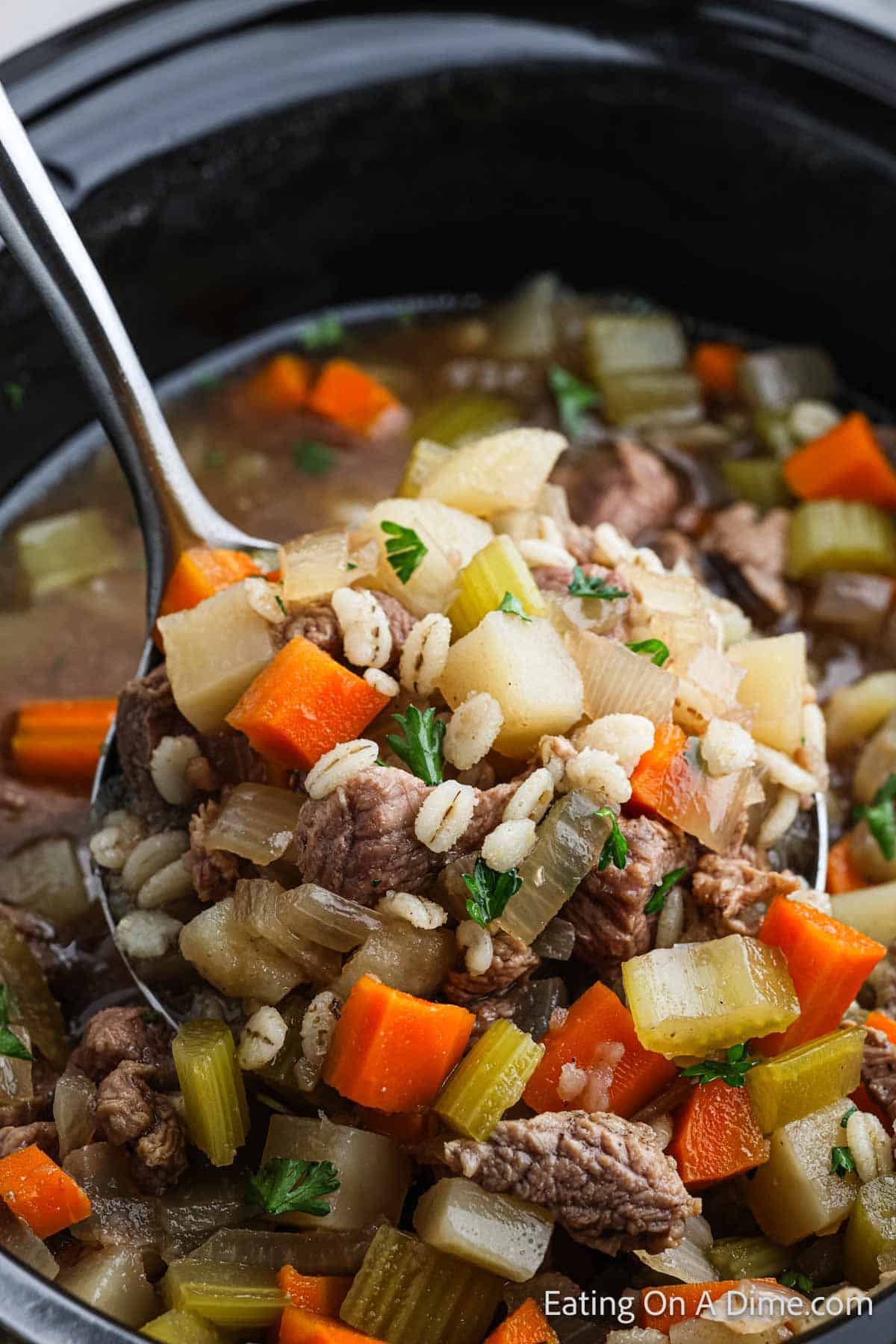 A hearty crockpot beef barley soup is shown in a ladle over a pot. The soup contains chunks of beef, carrots, celery, onions, potatoes, and barley, garnished with chopped parsley. The background reveals the rest of the savory concoction simmering in a slow cooker.