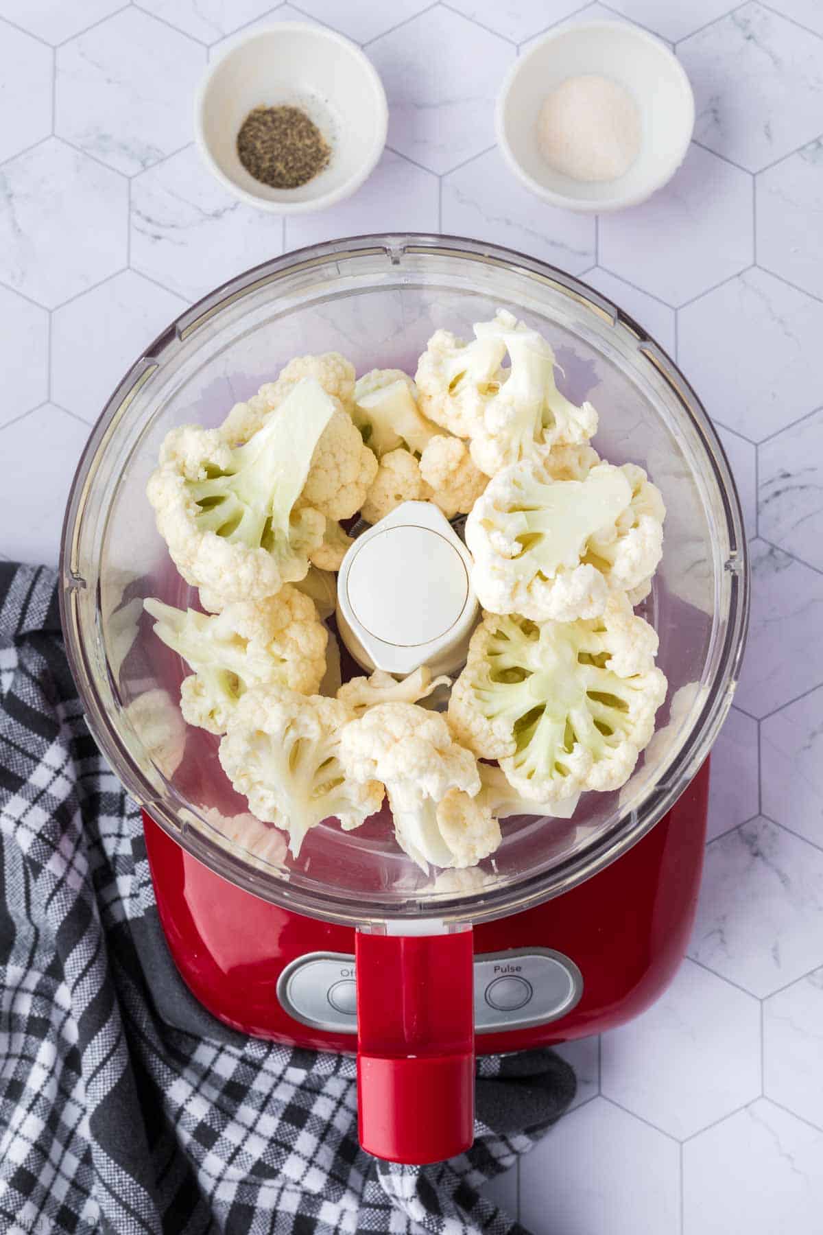 Chunks of cauliflower in a food processor with small bowl of pepper and garlic salt