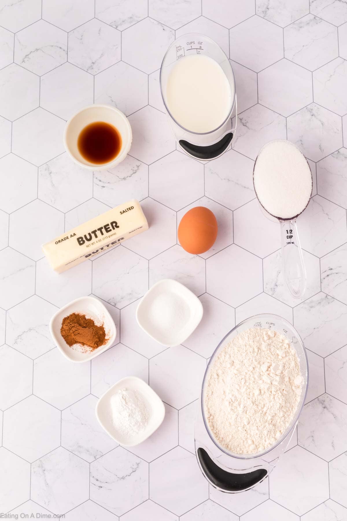 Top-down view of coffee cake recipe ingredients on a hexagonal patterned surface. Includes milk, vanilla extract, granulated sugar, a stick of butter, an egg, flour, cinnamon, baking powder, and salt. Measuring cups and a spoon are visible.