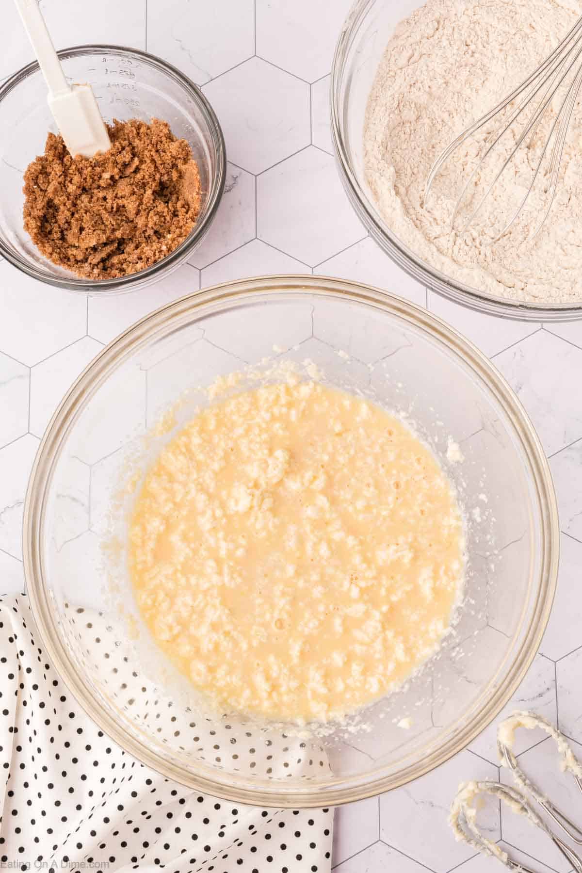 A baking setup for a coffee cake recipe features three glass bowls: one with a wet mixture, one with brown sugar and a spatula, and another with flour and a whisk. A polka-dotted cloth and electric mixer beaters rest on the hexagonal-patterned surface.